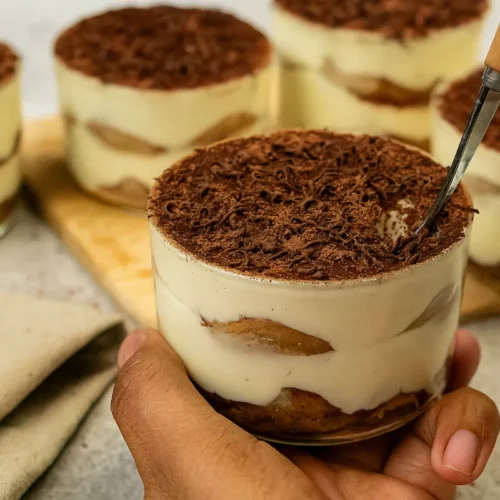 Tiramisu do pohára vrstvené s piškótami a krémom, bohato posypané kakaom, na drevenom stole.